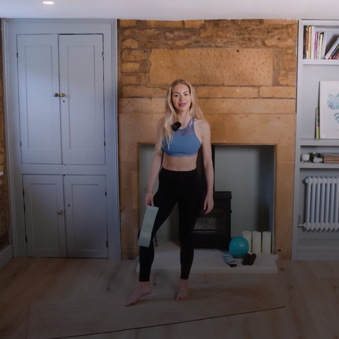 Woman in athletic wear standing in a room with a fireplace and wooden floor. amp wellbeing work out series for toning and energising