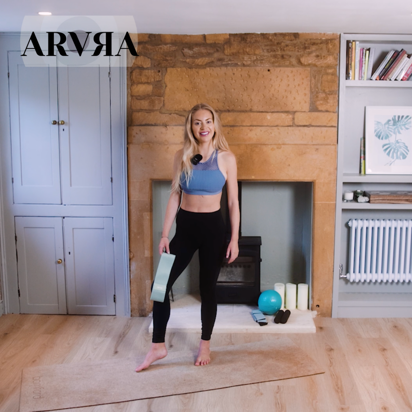 Woman in athletic wear standing in a room with wooden floor and fireplace, holding a yoga mat.