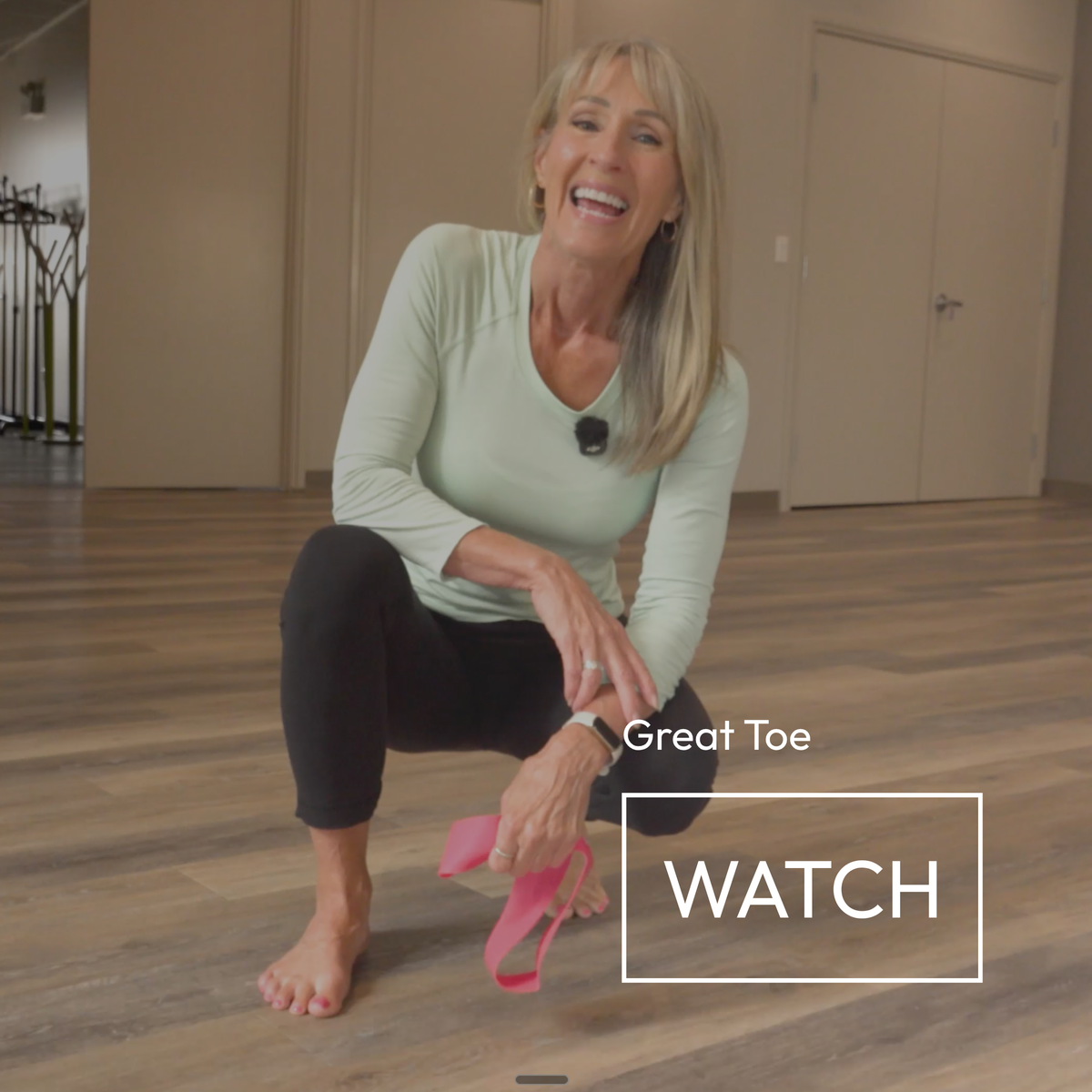 A woman crouches on a wood floor in a studio setting, demonstrating how to use a pink arch support band for her great toe. She is wearing a light sage green long-sleeve top and black leggings.