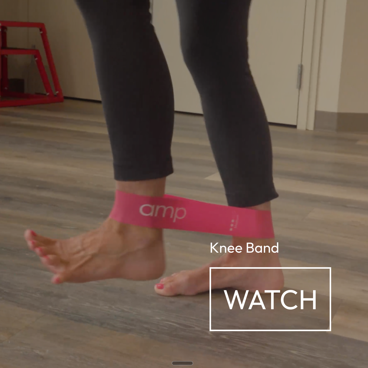 A pink Amp Knee Band worn around a person's ankle, shown in a home setting with wood floors. The image includes a 'WATCH' overlay badge.