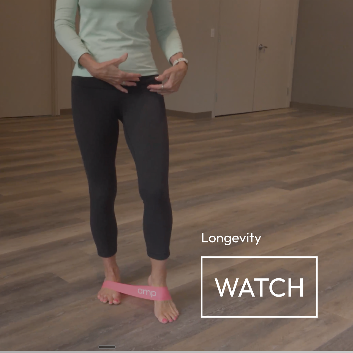 A person standing on a wooden floor indoors, wearing black leggings and a pale mint long-sleeve top, with a pink resistance band around their feet. White text on the right reads 'Longevity WATCH'.