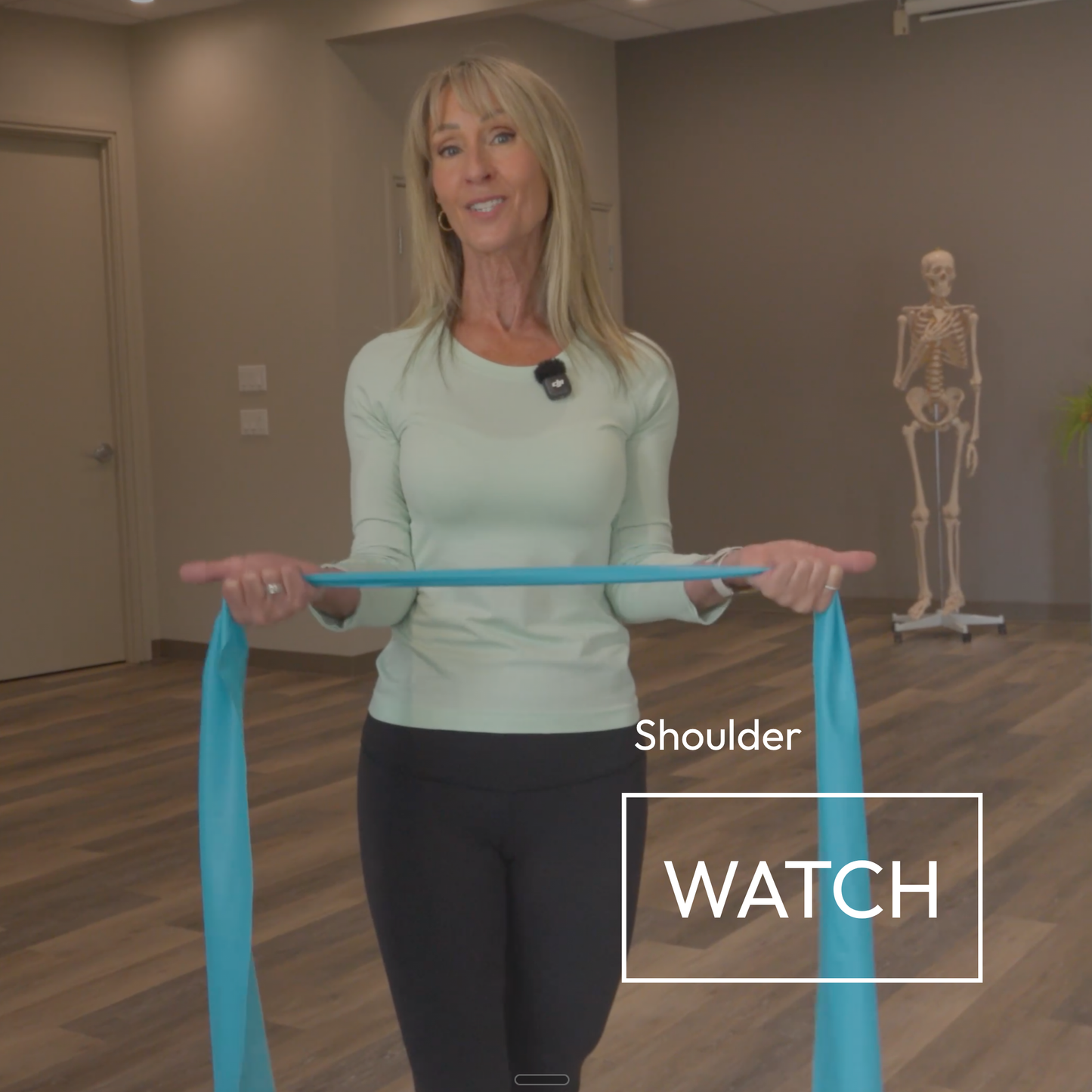 A woman in a light green long-sleeve top and black leggings holds a bright blue resistance band across her upper chest in an indoor studio. A skeleton model stands in the background, and text overlays read 'Shoulder' and 'WATCH'.