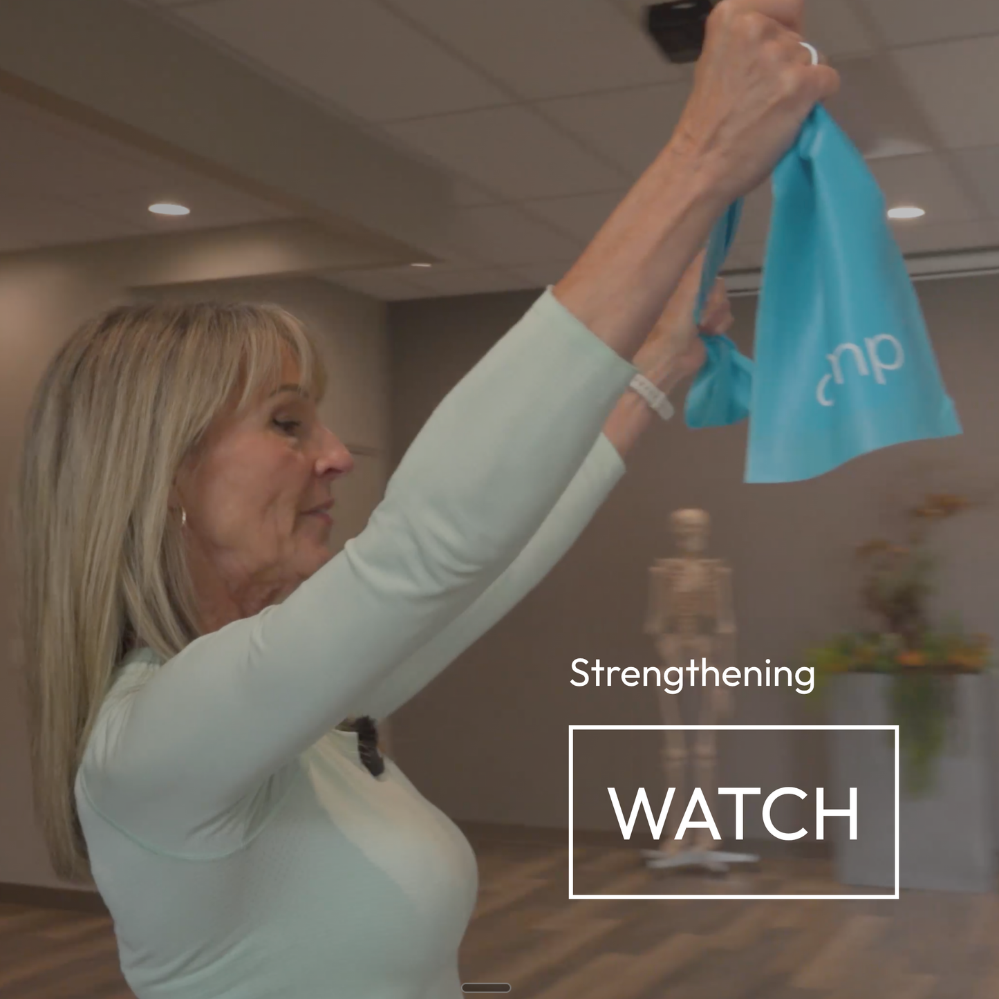 A woman in a pale mint green long-sleeve top holds up a light blue resistance band with the brand name amp. The background shows an indoor fitness studio with a mannequin and a 'Strengthening WATCH' text overlay.