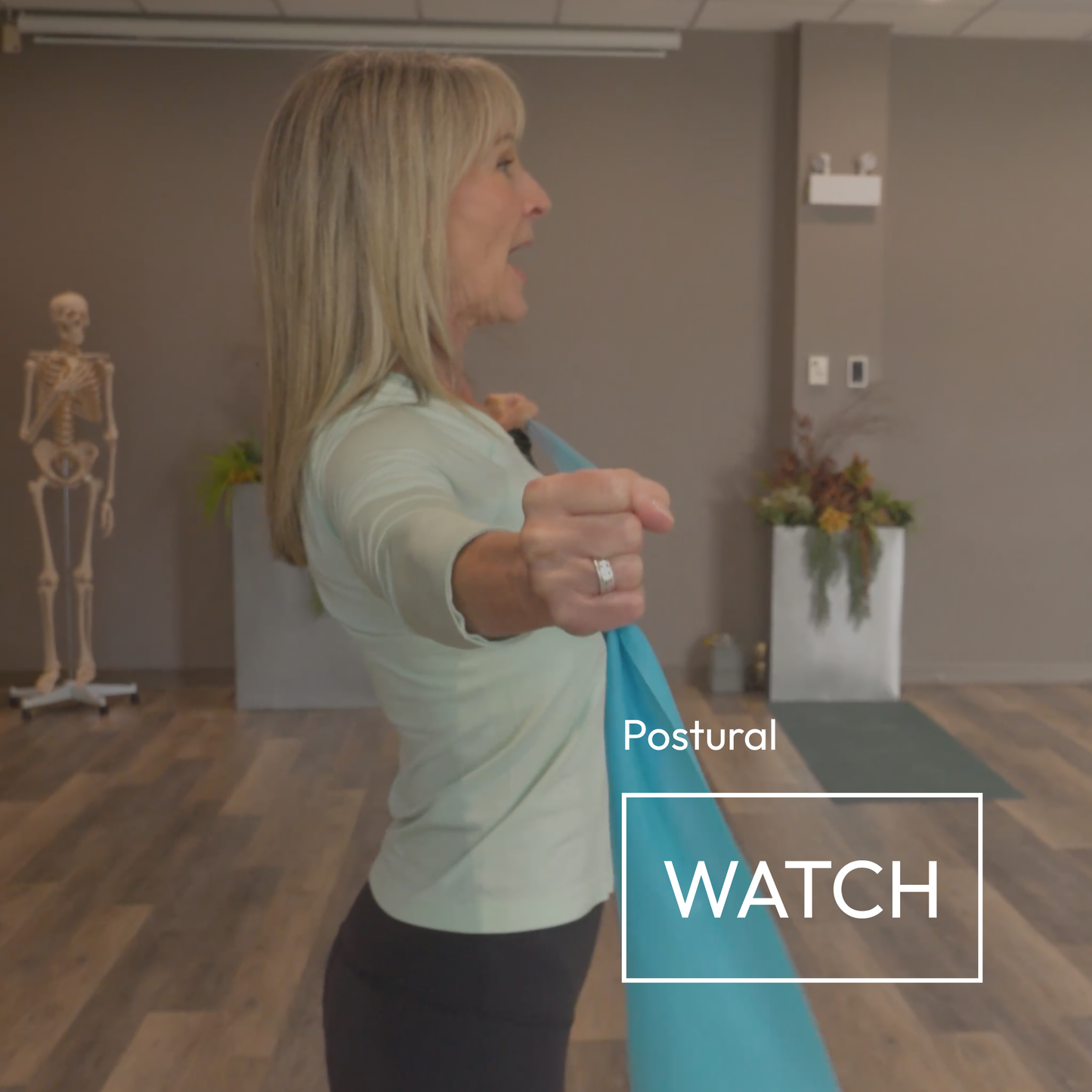 A woman in a light green top and black pants demonstrates a stretching pose using a bright blue yoga/exercise strap in a studio setting. The text 'Postural WATCH' is overlaid on the bottom right.